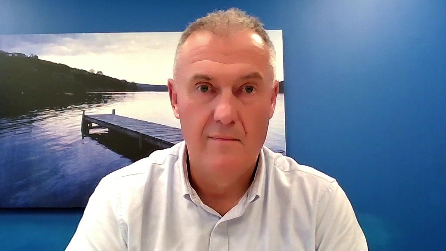 A head and shoulder shot of Dave Kaye looking at the camera. He has short grey hair and is wearing an off-white shirt. The wall behind him is painted a deep blue and features a canvas painting of a lake. 