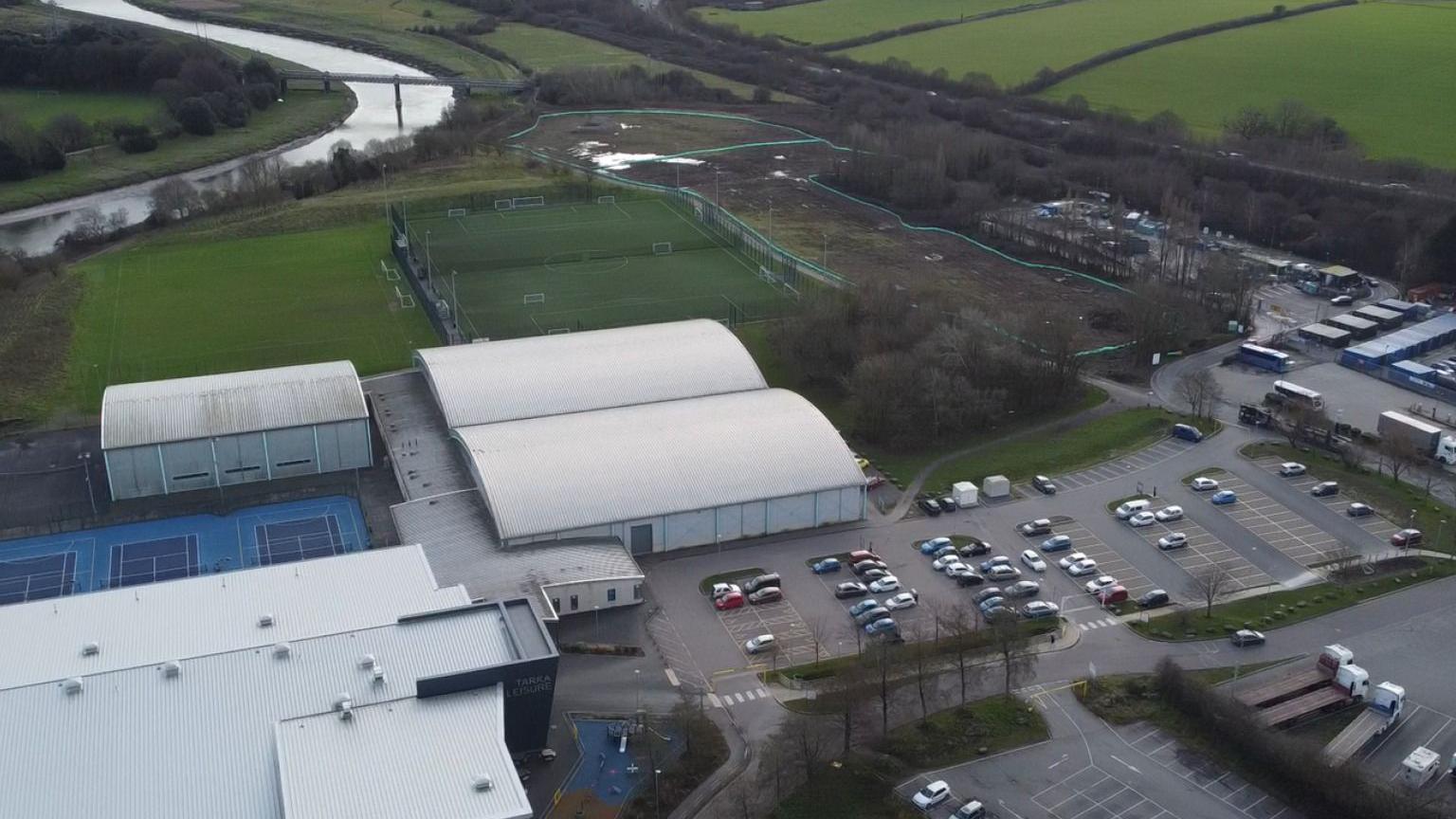 The Seven Brethren site for redevelopment from above. The top of the now demolished leisure centre is visible, as well as tennis courts and a football pitch. there is car parking and a road. The river is snaking off in the distance surrounded by green fields and countryside. 