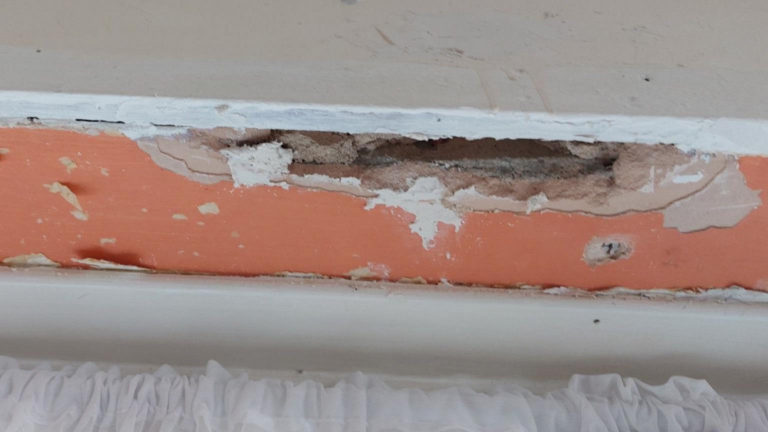Plasterboard with orange paint on a ceiling in a property. It has a large gaping hole where some has chipped away and looks in poor condition. There is a white curtain at the bottom of the image.