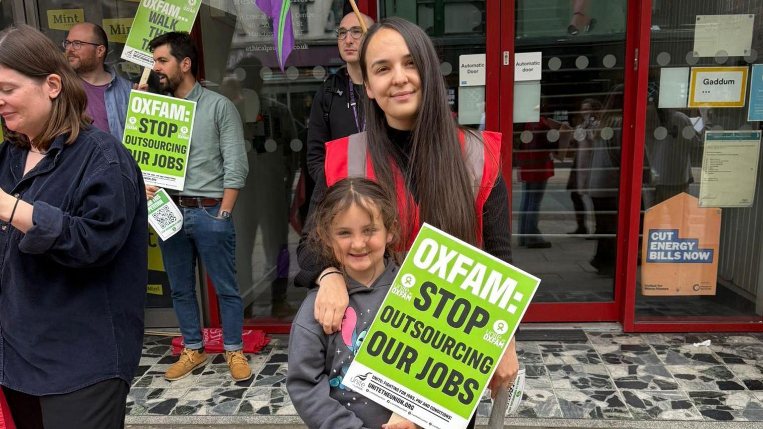 Protesters in a street. A woman has her arm around a young girl who holds up a placard.