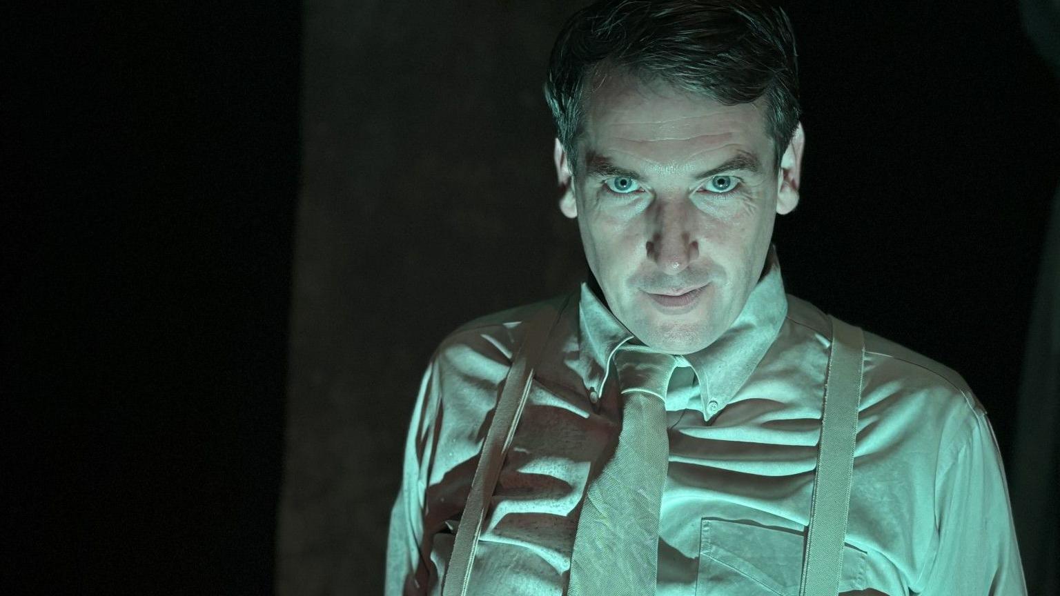 A man is staring at the camera. It is dimly lit and his clothing appears to be green