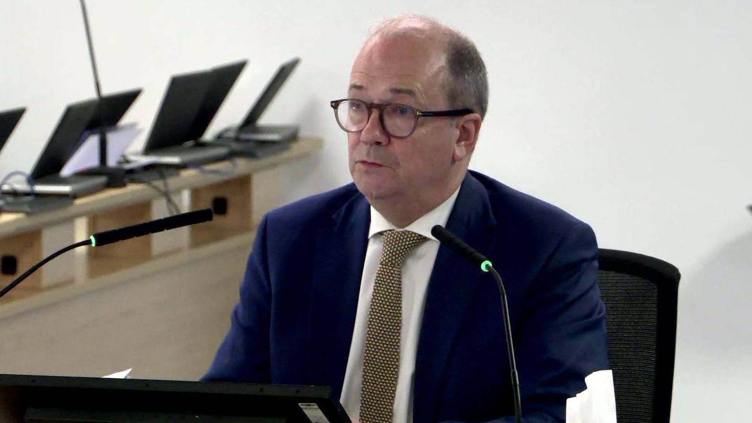 Dr Michael McBride is seated at a table with two microphones at either side of his mouth as he takes questions at the Covid Inquiry. He is balding and is wearing a pair of dark circular glasses. He is wearing a navy suit jacket, a yellow patterned tie and a white collared shirt.