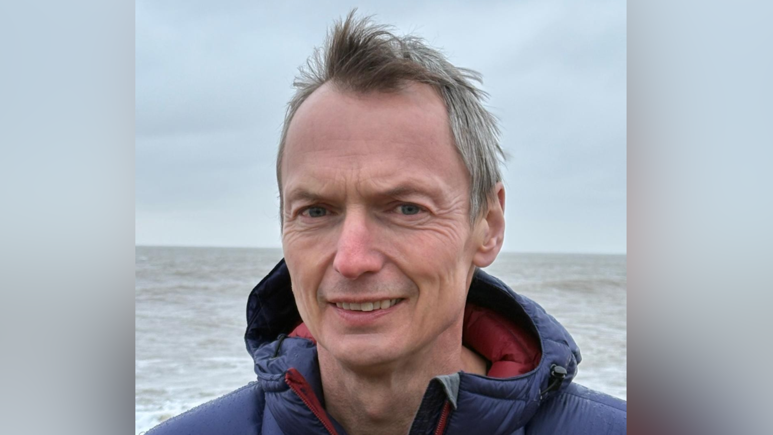 Hamish Ogilvie smiles at the camera as he stands on the coastline on a grey day. The sea is behind him. He has short grey hair and wears a blue rain coat.
