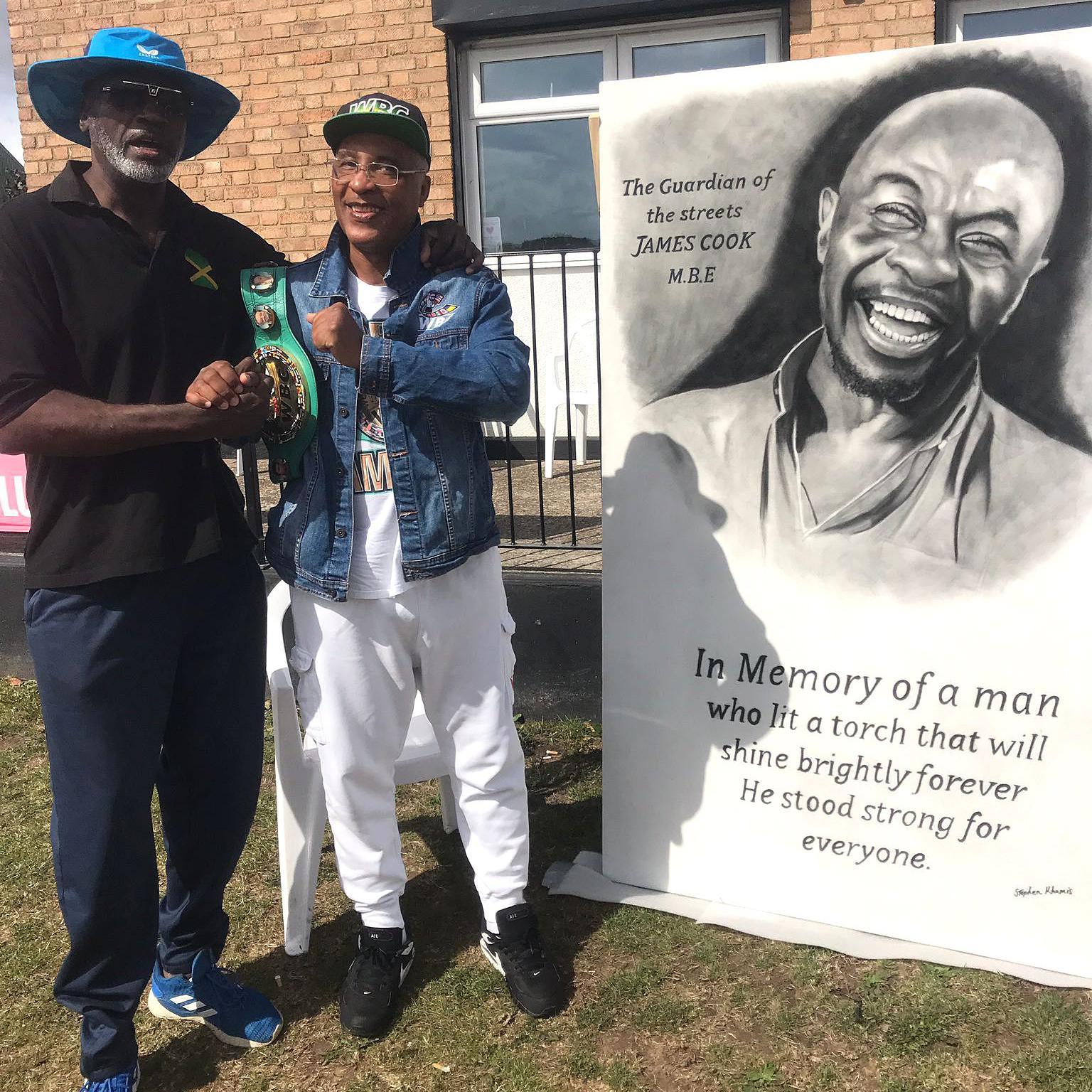 Former boxer Derek Williams with his arm around Michael Watson as they both stand next to a memorial painting of James Cook which reads "The Guardian of the Streets, James Cook MBE - In memory of a man who lit a torch that will shine brightly forever. He stood strong for everyone" 