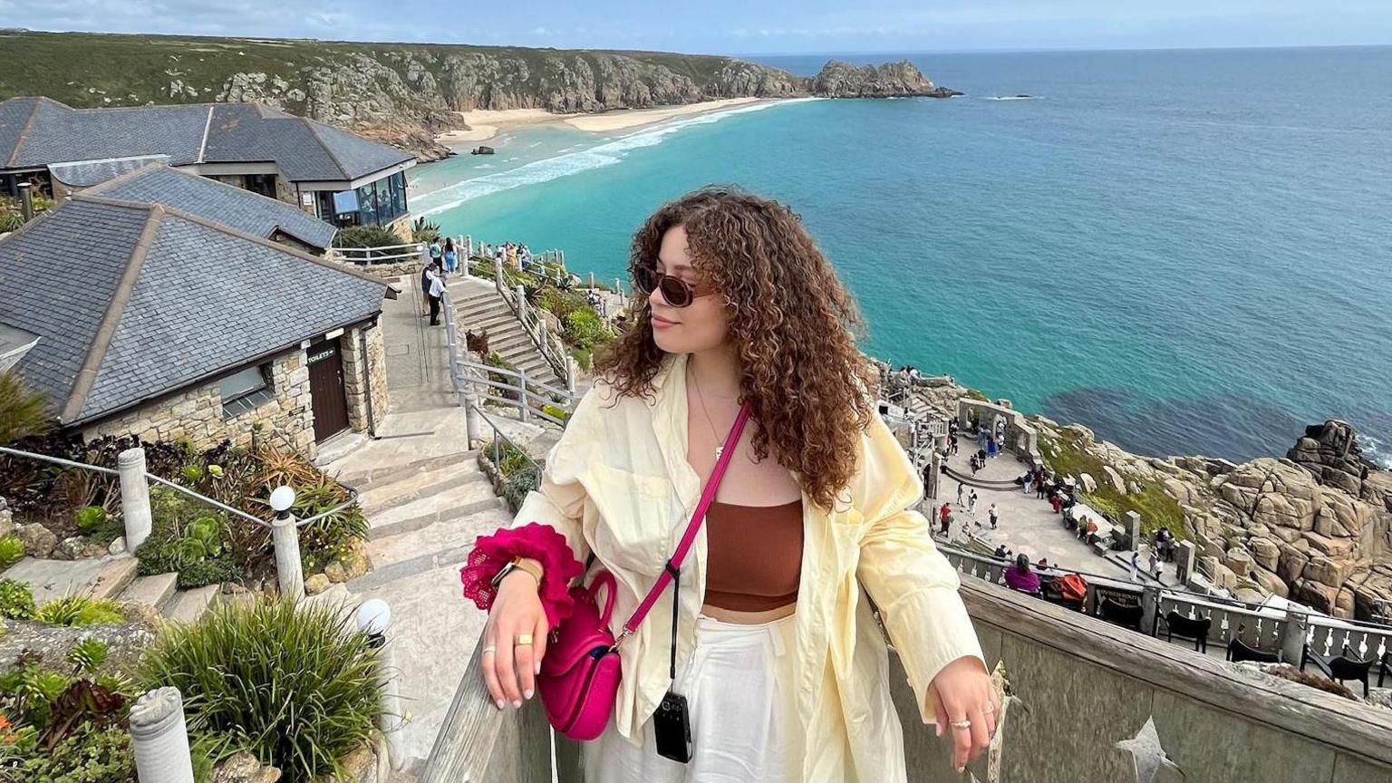 Eboni a woman in her 30s with curly brown hair standing above The Minack Theatre which is carved into the cliff face in Cornwall and covered in luscious green gardens with a view across turquoise sea
