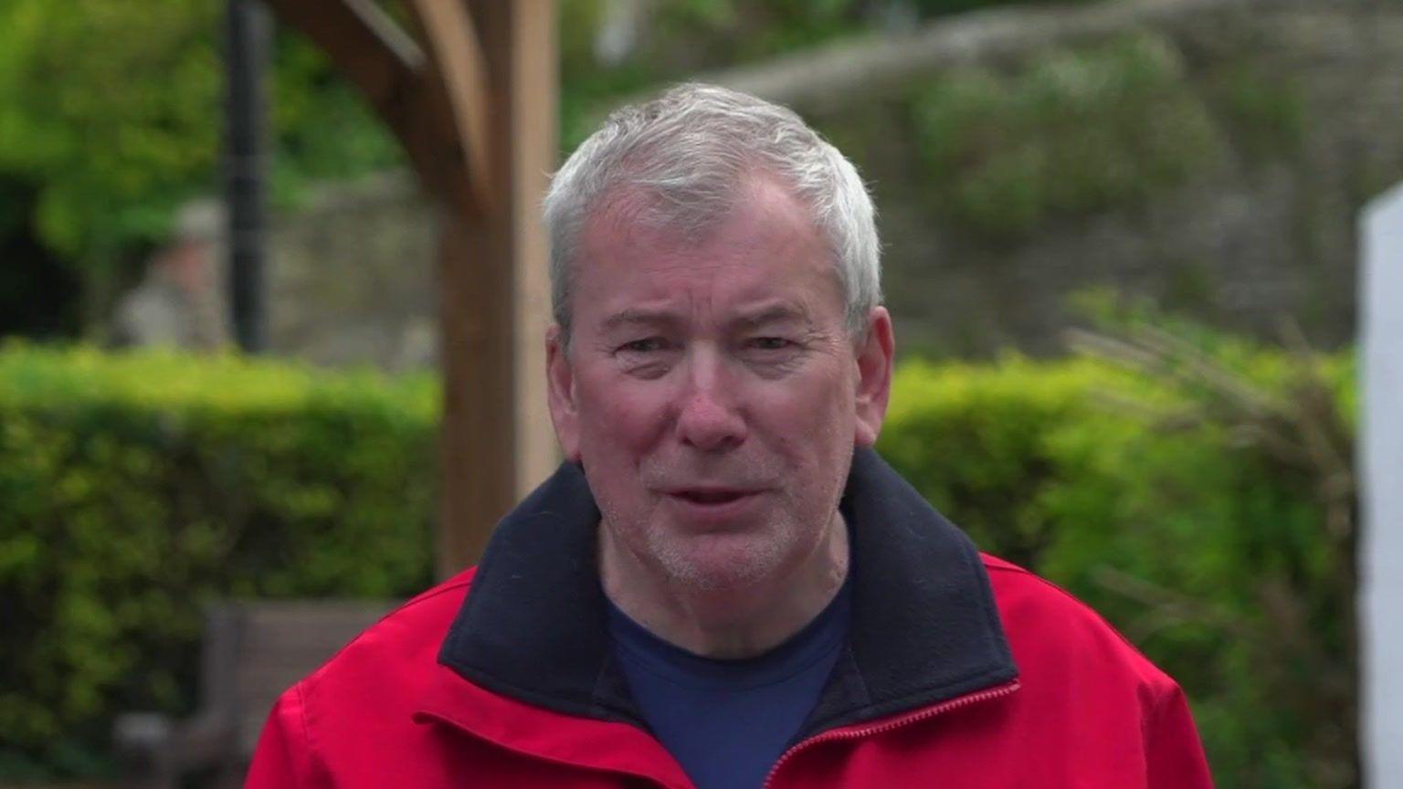 Malcolm Bell who is wearing a red coat and is standing in front of a hedge