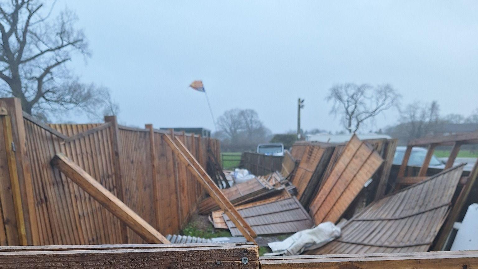 Brown fence panels on their side and on the floor after high winds, with trees in the background. 