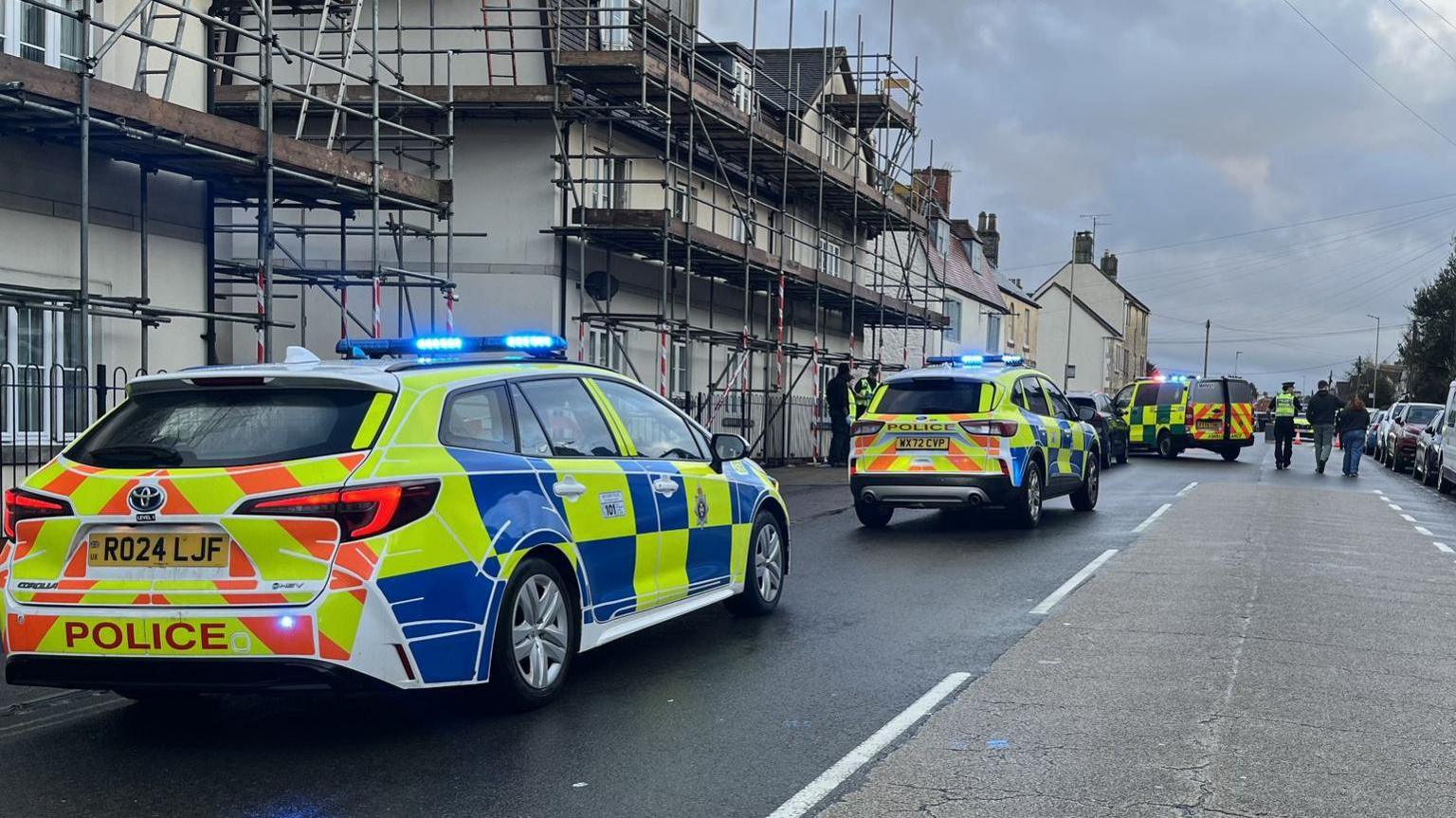 A road in Chippenham with houses on either side and cars parked. There are three police cars with sirens that are parked on the road.