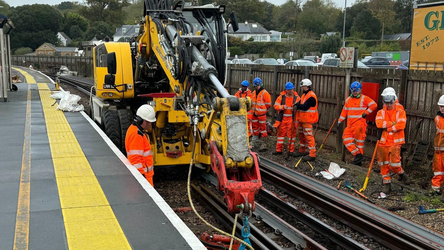 A large, wheeled machine on a railway deploys a crane-like projecting arm to drag track into place. Nine railway workers wearing helmets and orange overalls stand beside a railway station platform.