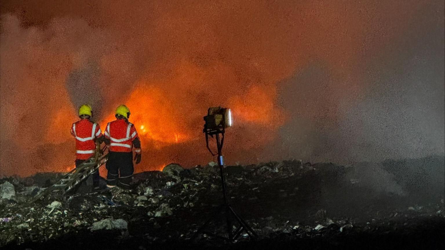 Two firefighters watch on as fire burns at a landfill site. They wear yellow helmets and orange high-vis jackets.