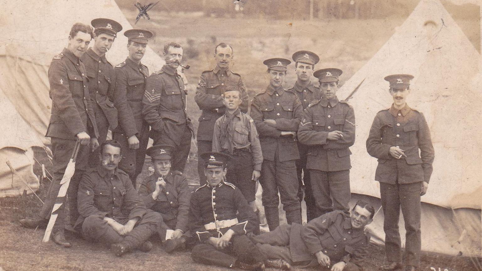 A black and white photo shows 13 soldiers in uniform and a boy - standing and sitting in front of tents. They are dressed in uniform. An "X" is written above the soldier third from the left and on the back it is said to indicate this is Rupert Thompson.