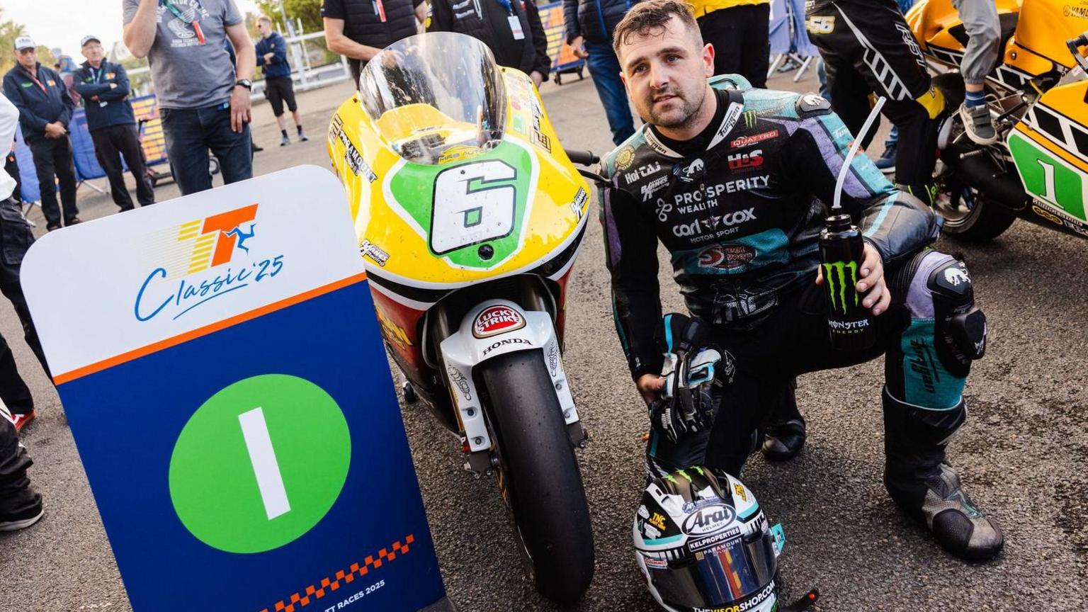 Sunlop kneeling next to the bike, which is yellow at the front with a white number six on a green plate. There is a Classic TT board with the number one on it next to the bike.