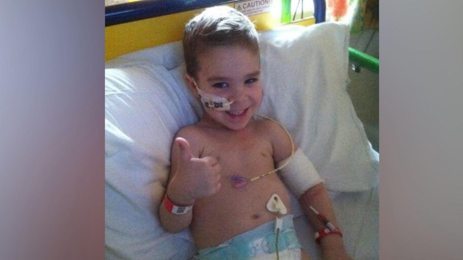 Finley as a young boy, lying in a hospital bed with tubes attached to his body, arm and face. He is wearing a nappy. He is smiling at the camera and giving a thumbs-up with his right hand.
