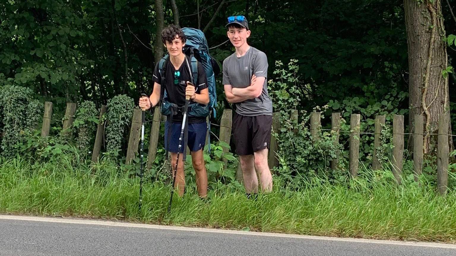 James McCarthy-Hill and Adam Mitchell standing on a grassy verge beside the side of a road. They are both wearing shorts and T-shirts. Adam has blue reflective sunglasses resting on his black cap, and James is carrying a large blue backpack and holding two hiking poles. Behind them there is a log and wire fence and thick woodland.