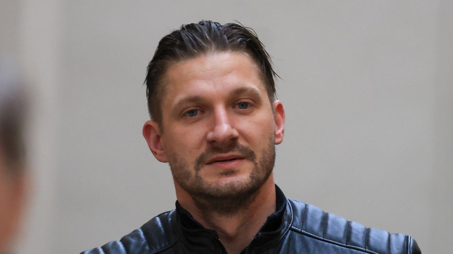 Karolis Peckauskas has short black hair and beard. And is wearing a black jacket.