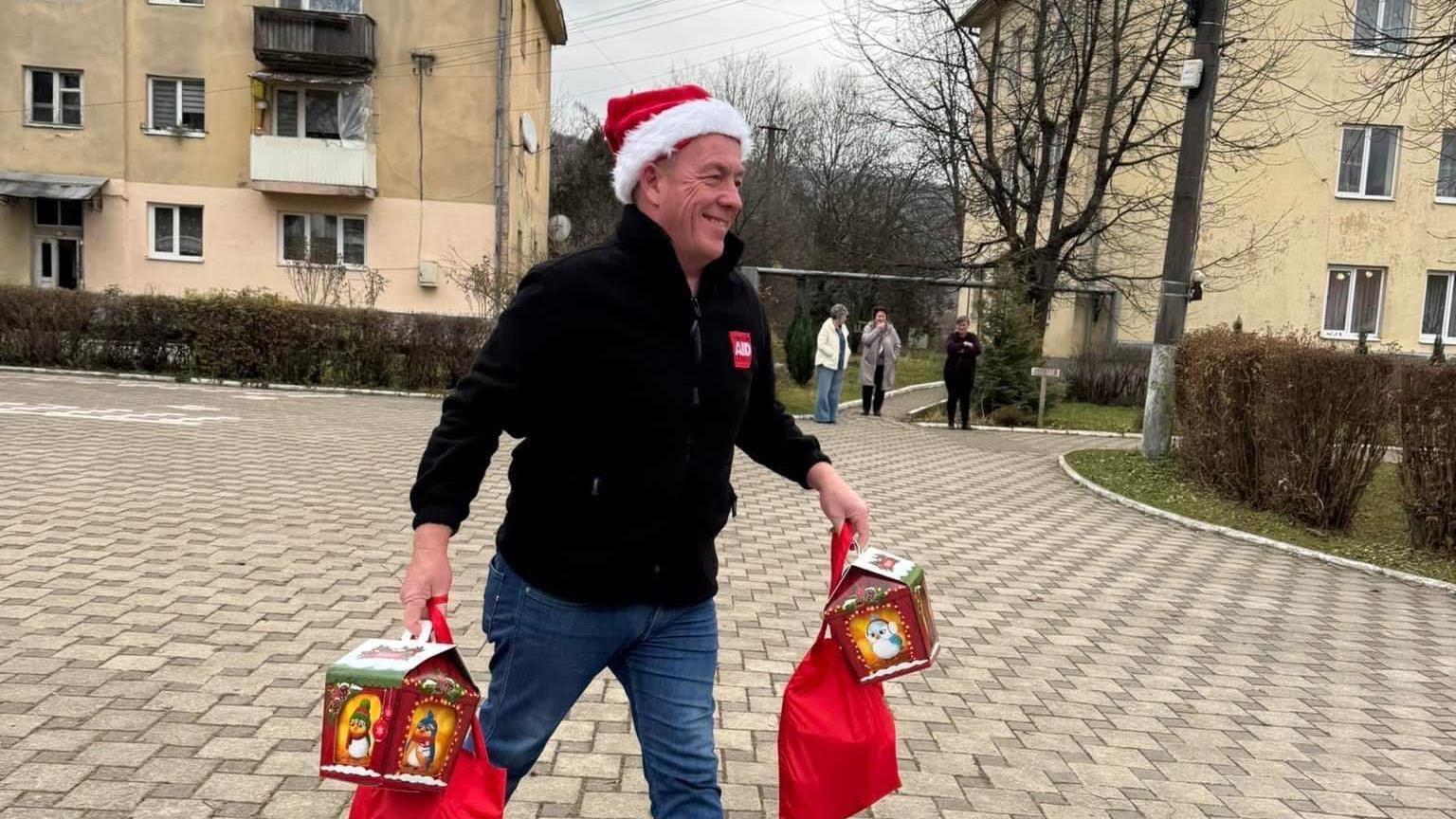 A man in a black fleece and jeans, wearing a Santa hat, walks through a residential area carrying gifts.
