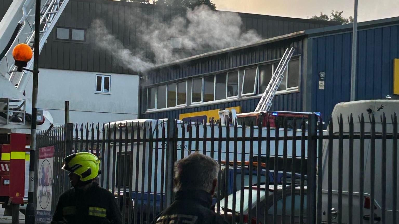 A blue industrial building with smoke coming from the top. Two firefighters at the front of the image, one on left wearing a yellow helmet. 