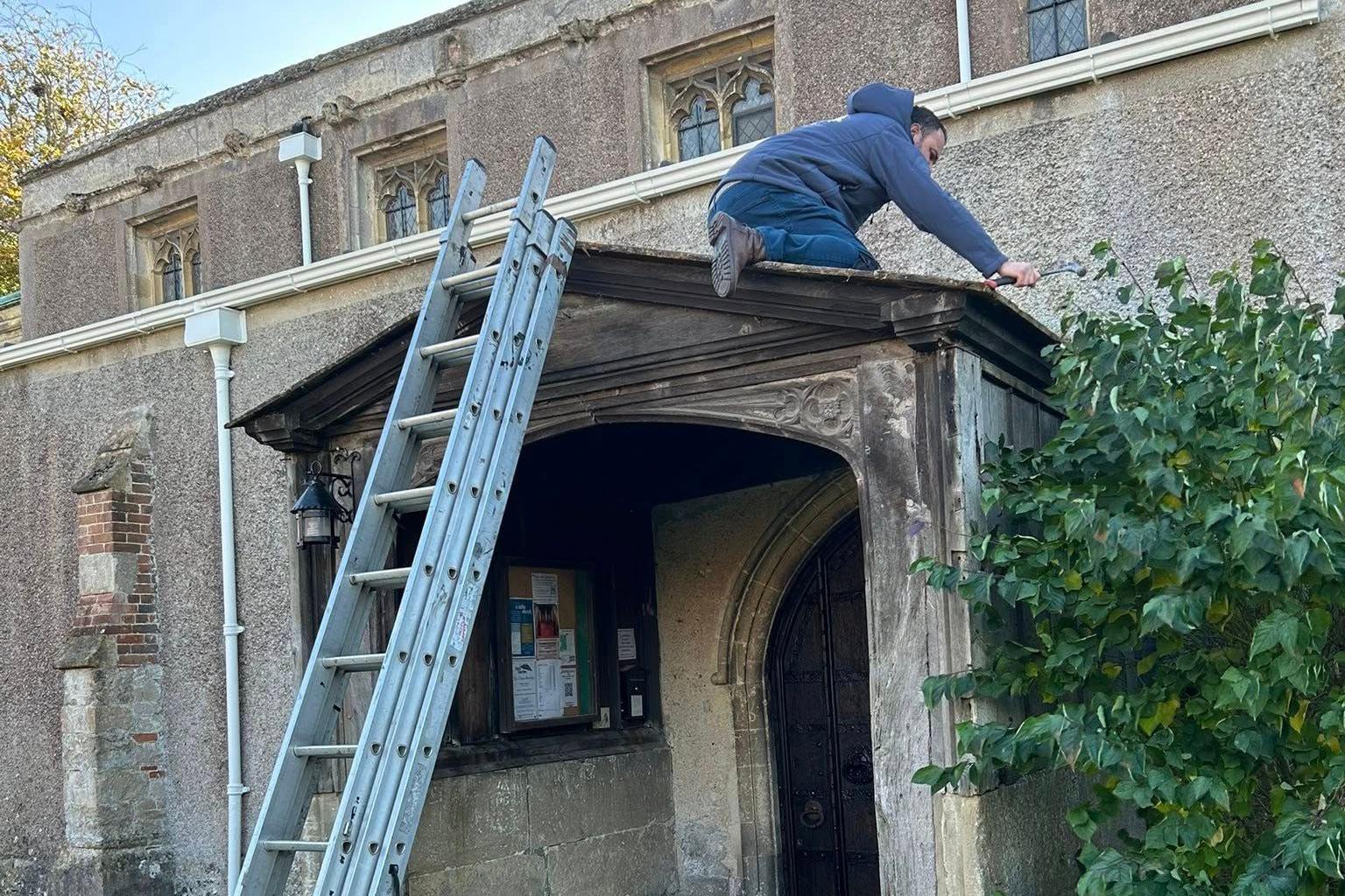 Michael is on the roof of the porch with his tools, a metal ladder leaning against the structure. Inside the porch a noticeboard and a wooden door are visible.