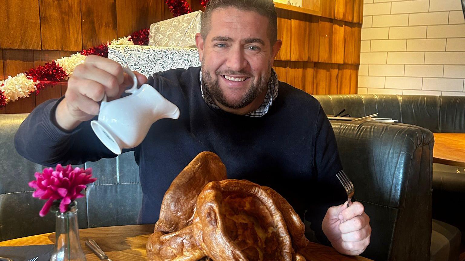 A man pouring gravy onto a mound of large Yorkshire puddings.