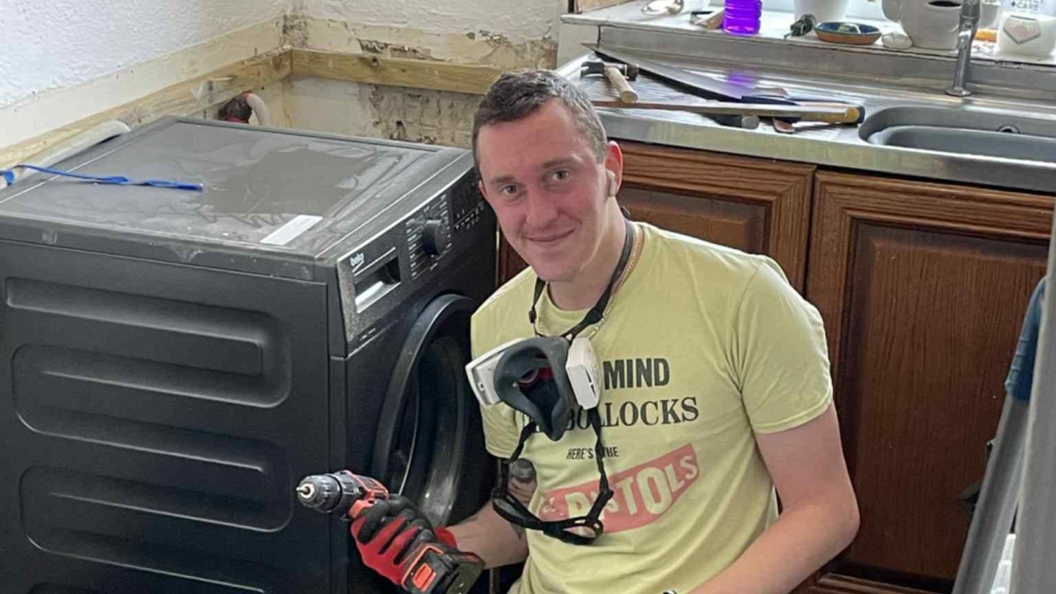 A man sitting by a washing machine with a drill in his hand, carrying out repairs. He is wearing a Sex Pistols shirt.