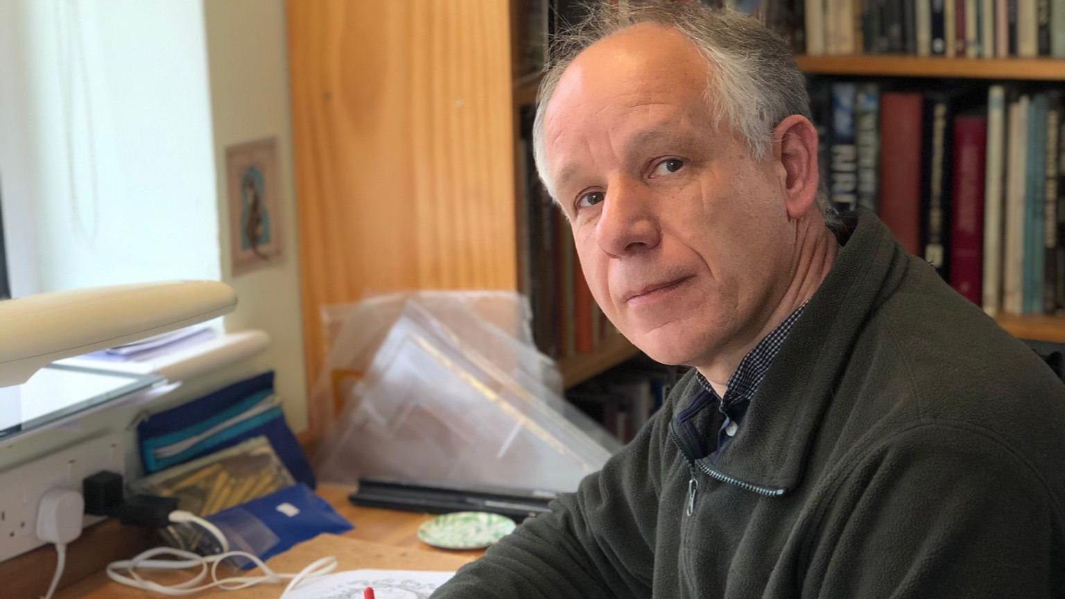 Tim Noad sits at his desk - he is looking at the camera from the right, and wears a dark fleece. There's a bookcase in the background.