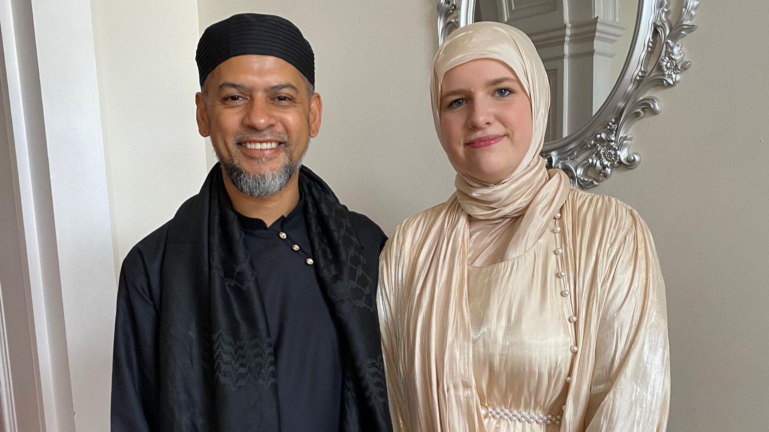 Mohammed Mokuzzal Ali with his daughter, Aleyah Knight. Mohammed is wearing all black. Aleyah is wearing a cream silk hijab and dress. They are stood side-by-side, looking at the camera and smiling.