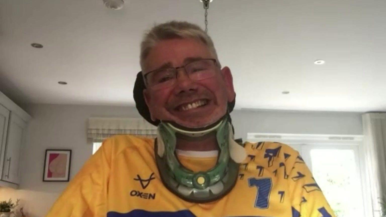 A man with white hair and glasses wearing a neck brace and a yellow and blue T shirt with number seven on. 