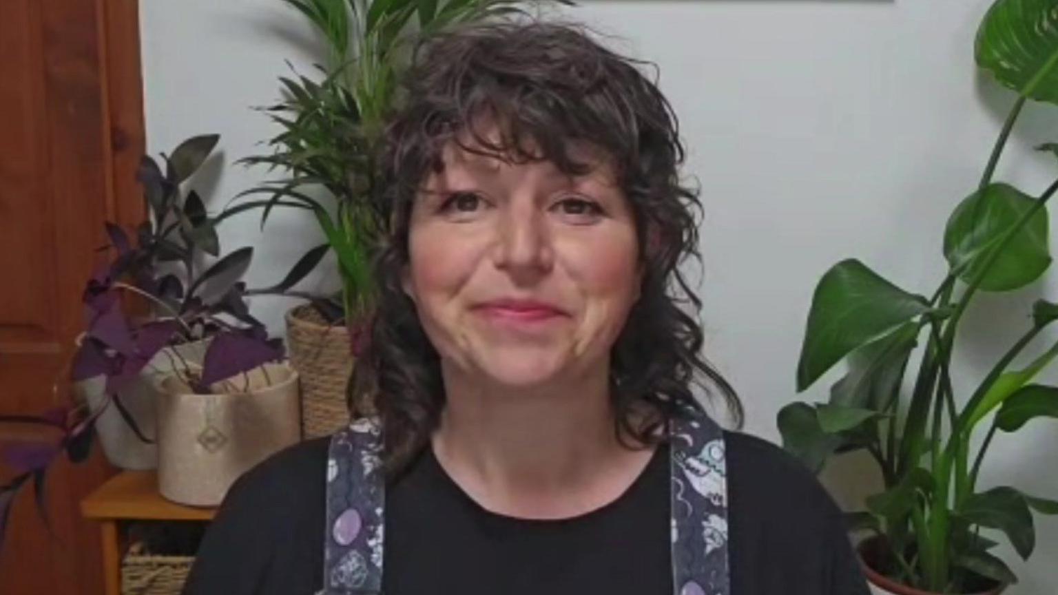 A woman on a video call with houseplans behind her. She is smiling and has curly brown hair. She is wearing a black top and what looks like purple and grey dungarees.