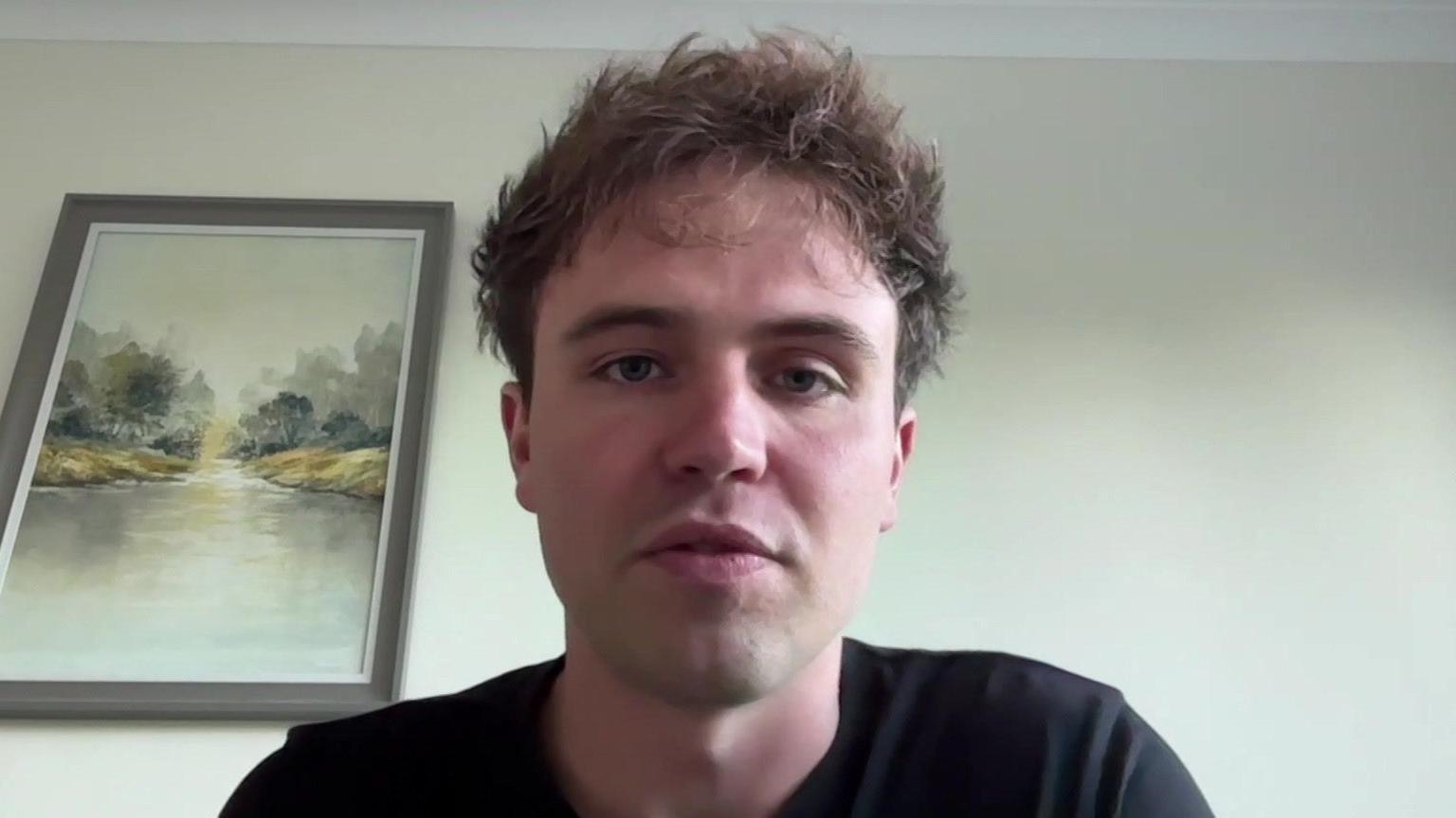Matt Staniek during an online interview. He is looking at the camera. He is wearing a black T-shirt and has short messily styled hair. A painting of a lake hangs on the wall behind him.