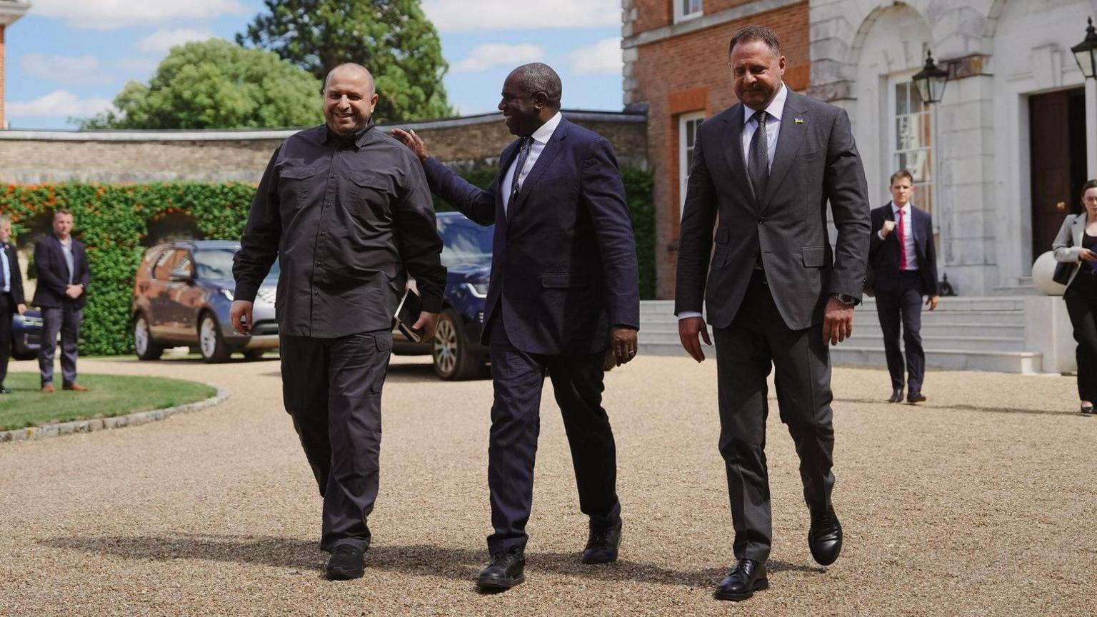David Lammy, wearing a suit, shirt and tie, is seen placing his arm on the shoulder of a smiling Rustem Umerov, who wears a dark military-style outfit, as Andriy Yermak, wearing a suit, shirt and tie, walks alongside them on a gravel driveway outside the front of Chevening, a large country house