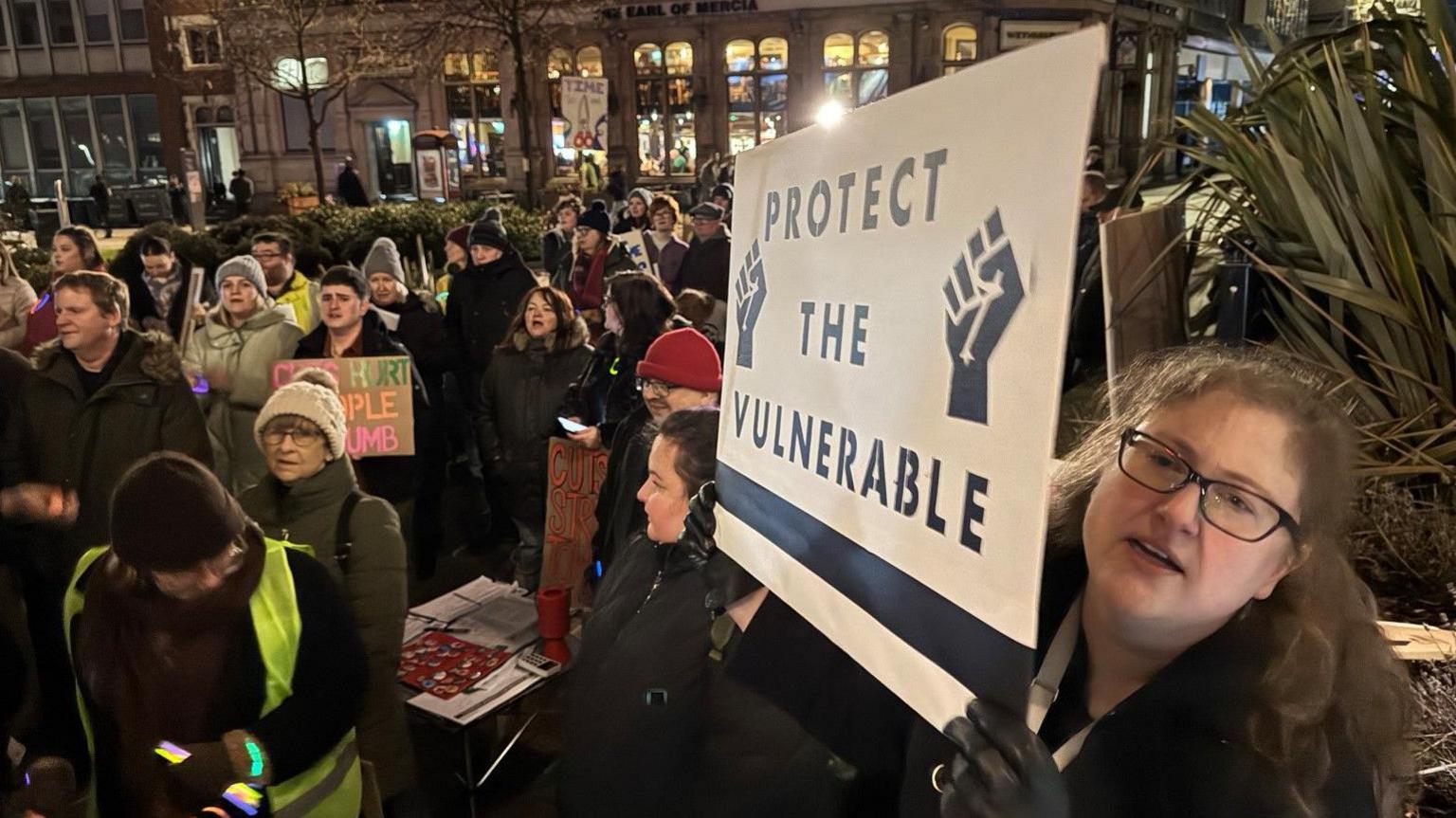 Person holding a banner in the foreground that says protect the vulnerable. Other people are in the background.