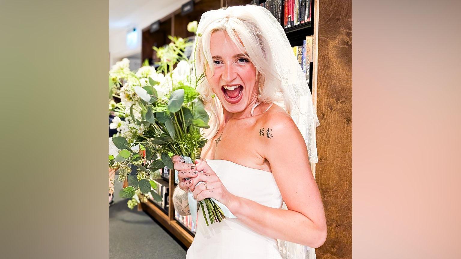 A woman with long blonde hair and wearing a white strapless wedding dress and white veil is smiling while holding a bouquet of flowers. She is standing in front of shelves of books as part of the Storyhouse library.
