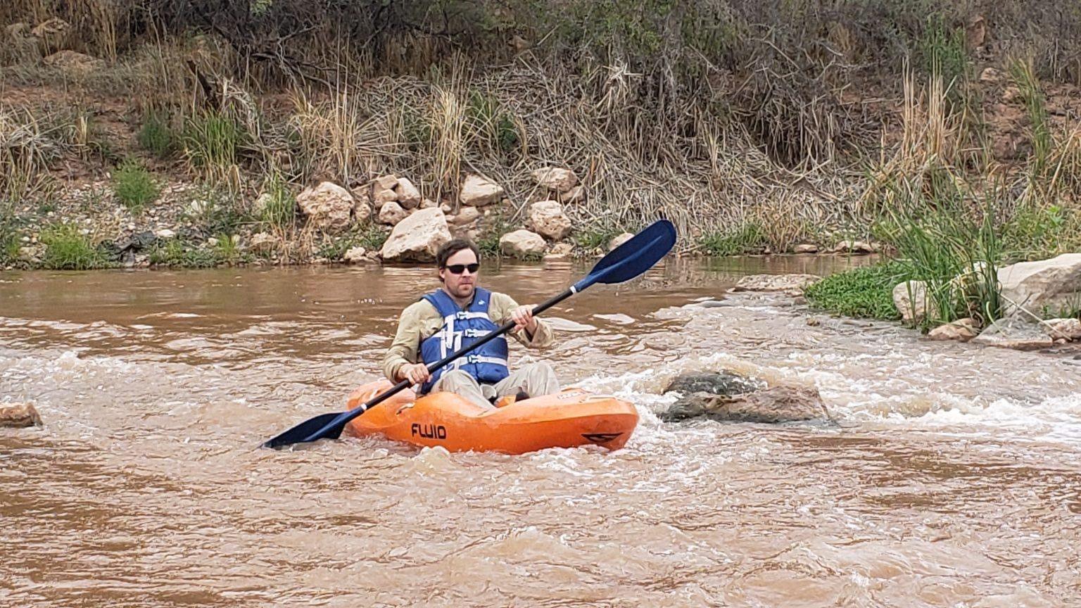 Mr Perrett is in an orange kayak in a river with greenery on the banks. He has brown hair and is wearing sunglasses. He has a blue life jacket on and is holding a blue double ended oar.