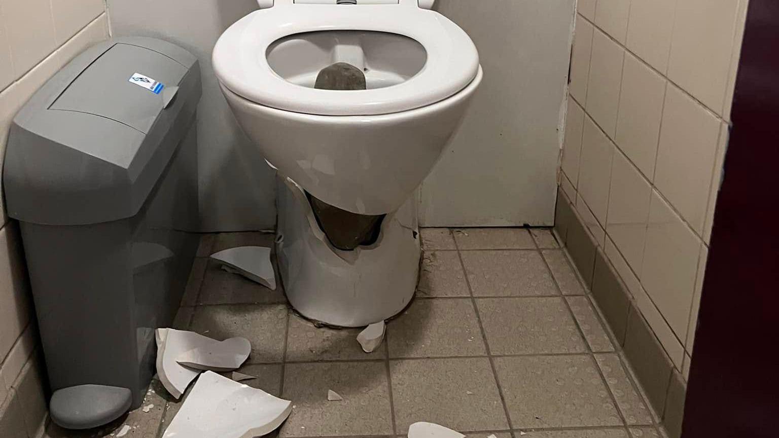 A broken toilet bowl in a small tiled public toilet. Large pieces of the broken bowl are on the floor along with smaller shards.