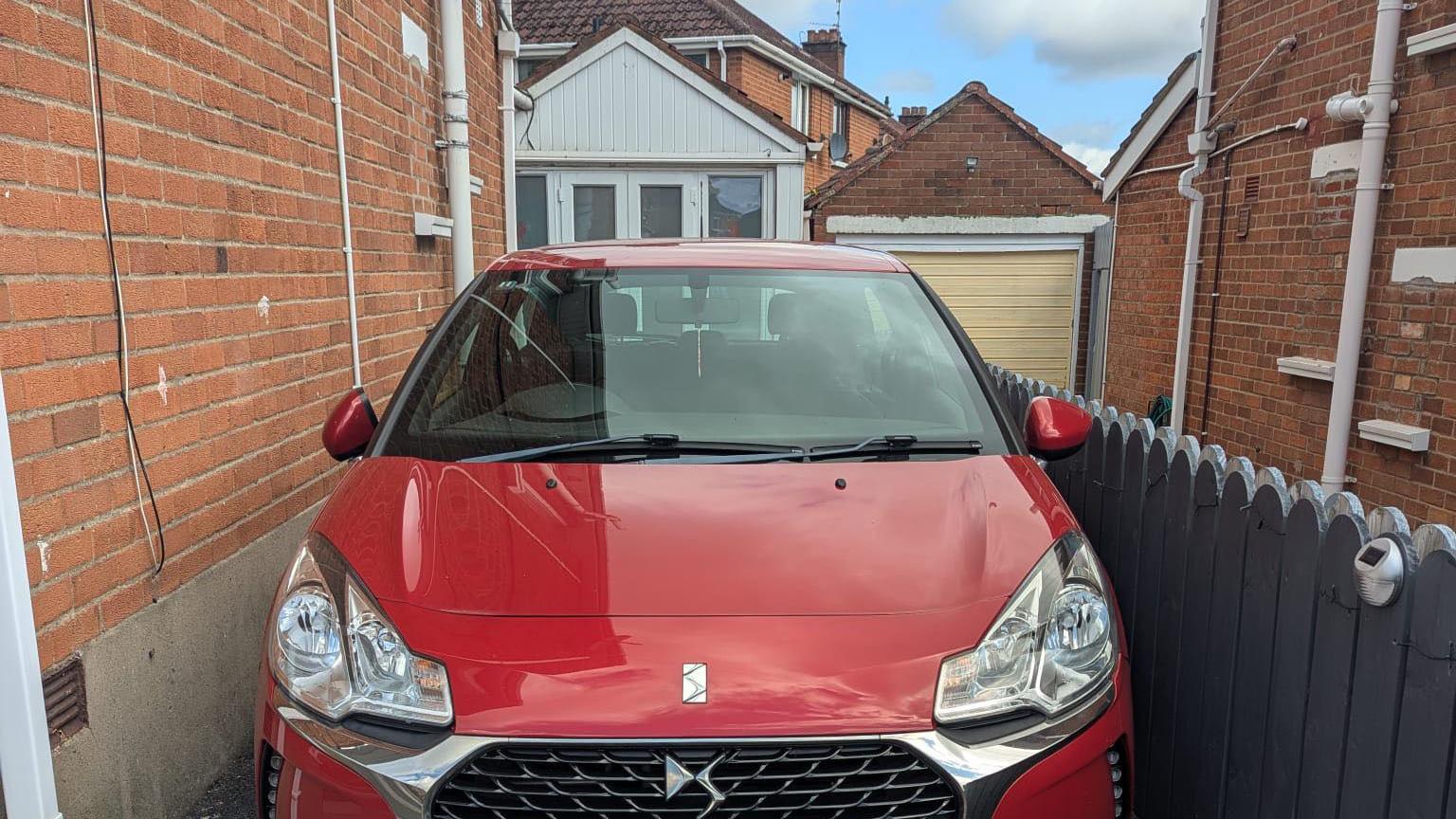 A red Citroen car sitting in a driveway with a grey gate on the left and brick wall on the right.