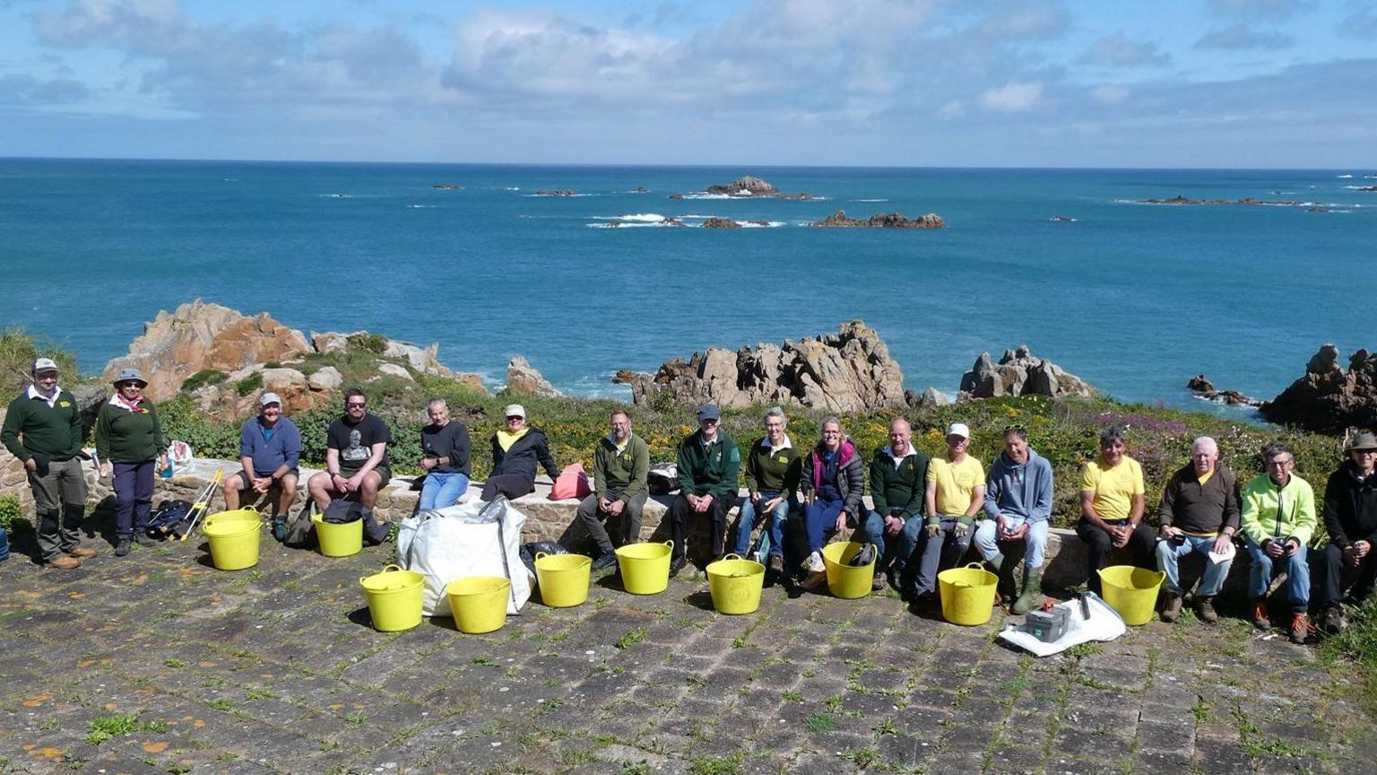 Image shows a large group of volunteers sitting on rocks, against a backdrop of the blue sea and coastline, with large yellow collection buckets in the foreground.