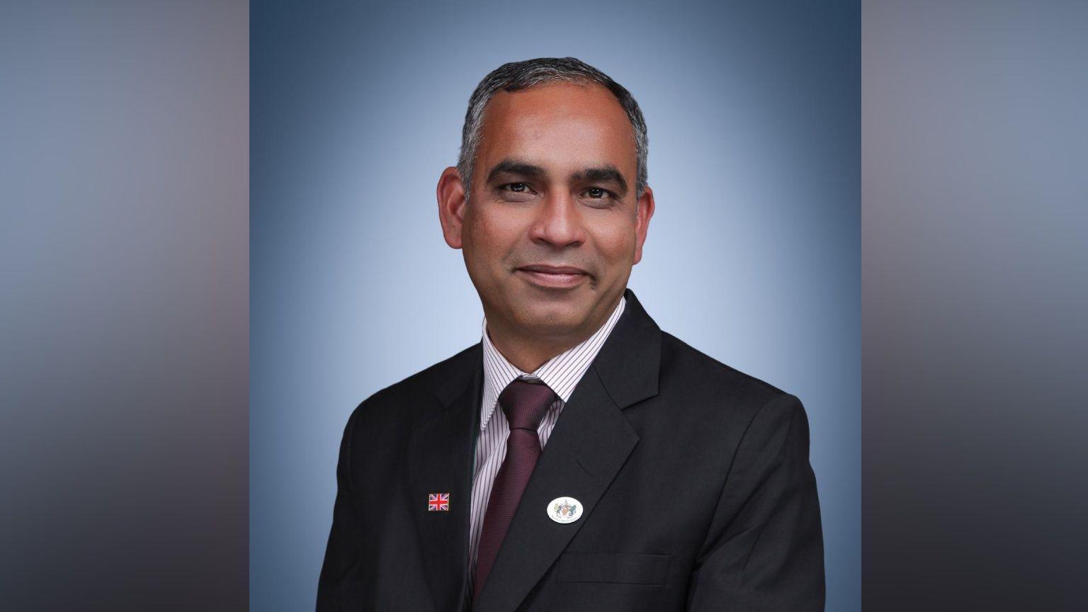 Adi Malviya wears a dark-coloured suit, striped shirt and purple tie. He faces the camera. He sits against a plain blue background.