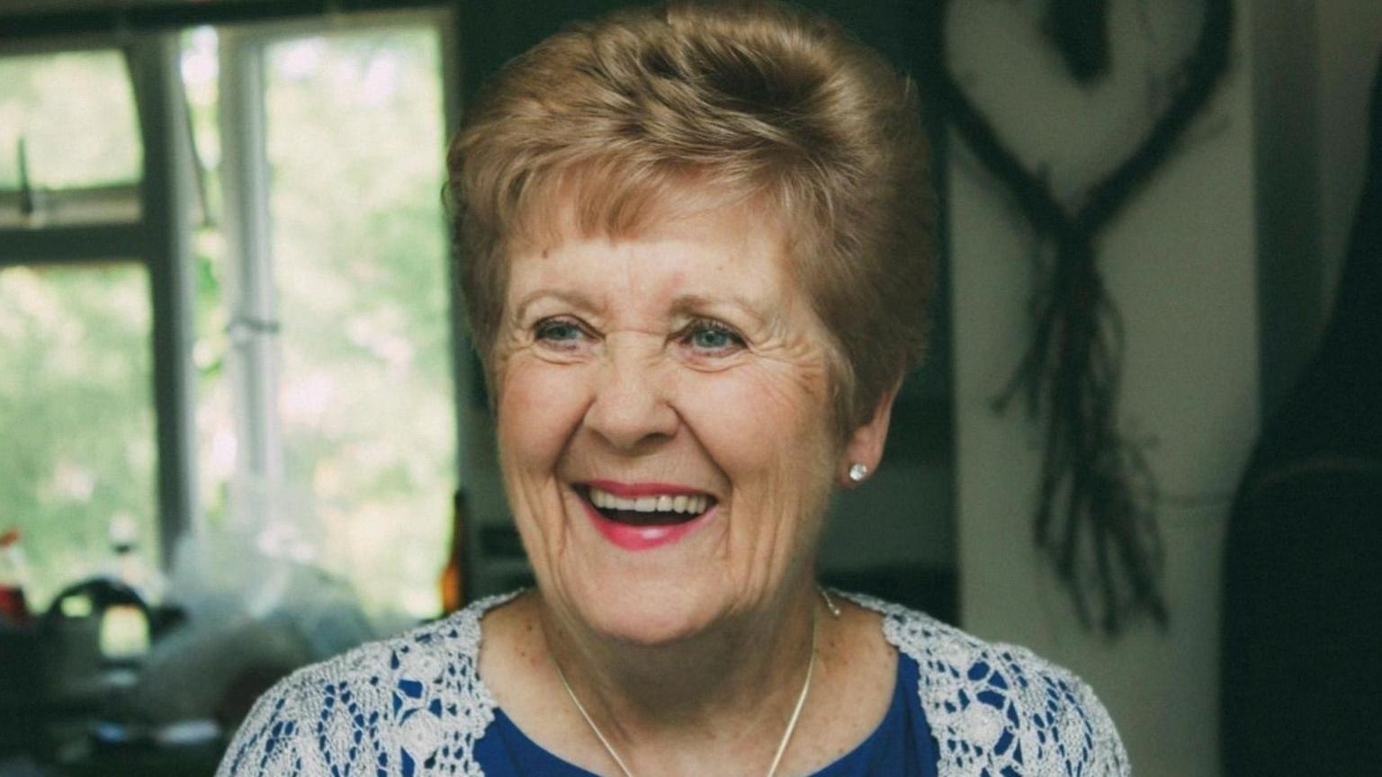 Beryl Howard wearing a blue top underneath a white lace cardigan, a silver necklace and bright pink lipstick. She is looking off to the left of the camera and laughing. She has short light brown hair and blue eyes.