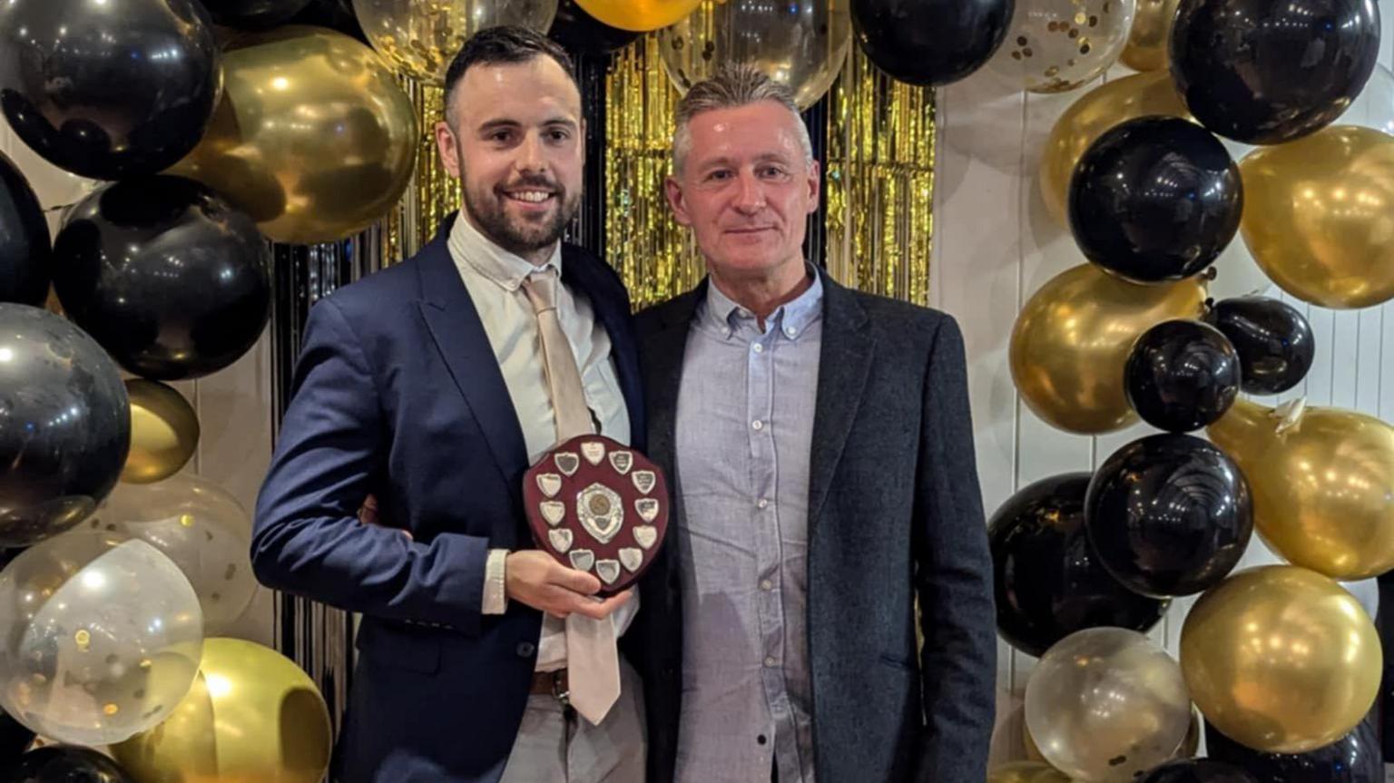 Lee and Darren pictured at an event with black, gold and transparent balloons seen behind them. They are wearing formal, suit attir3e and Lee is holding a plaque
