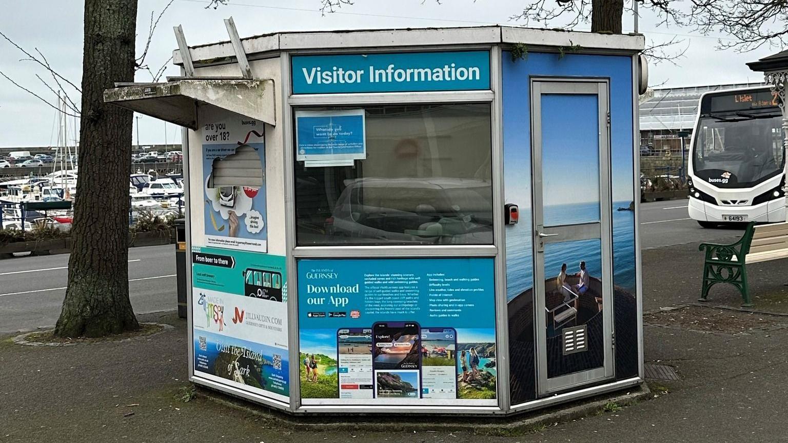 The hexagonal unit. Three sides of it can be seen. The left one has a covered hatch, the middle one has a window and the one on the right a metal door. There is a sign reading "visitor information" and there are tourism-related marketing posters covering the rest of it.