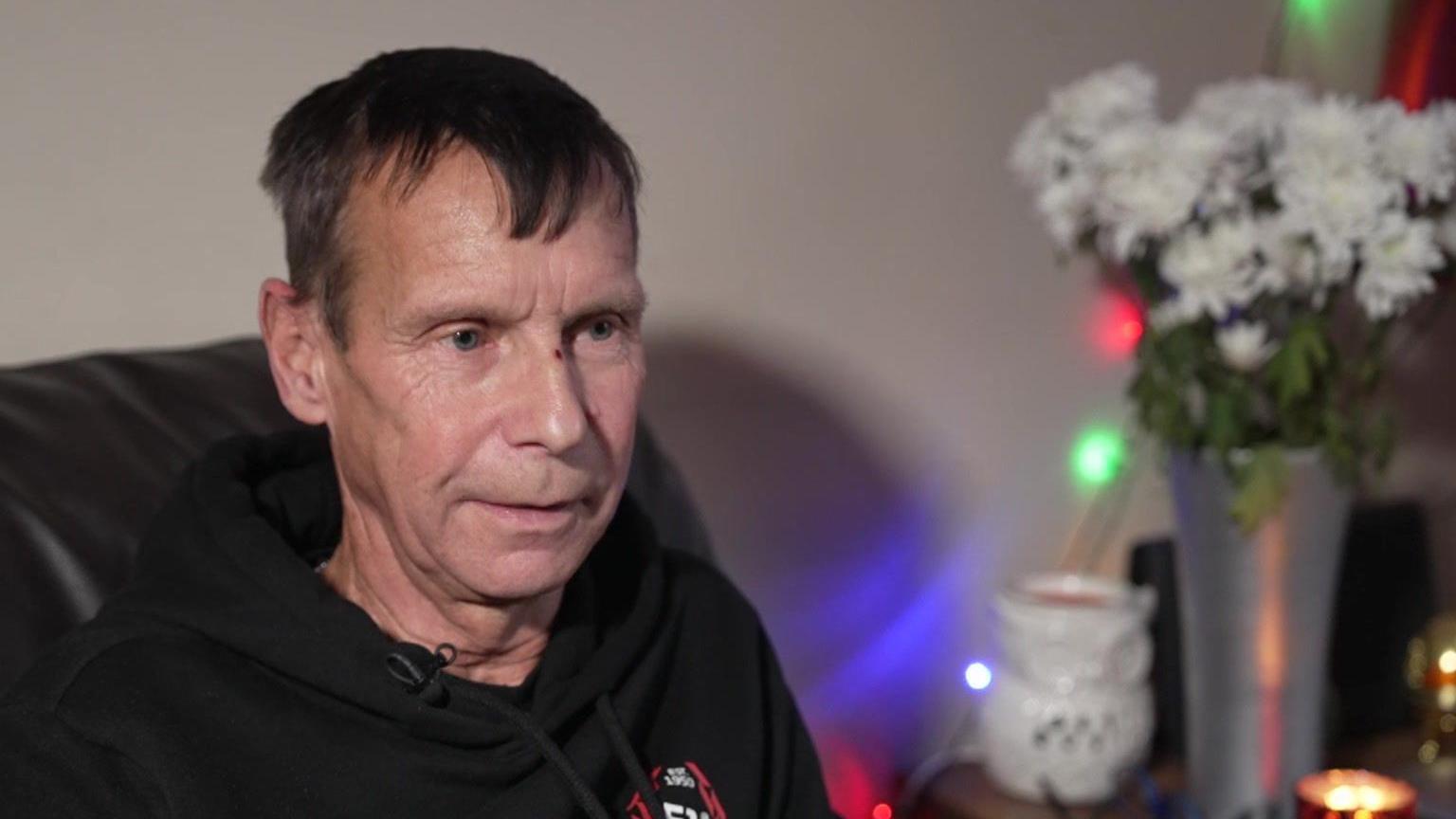 A man being interviewed on BBC TV sat on his own coach, with a bunch of flowers and a lit candle on the side table