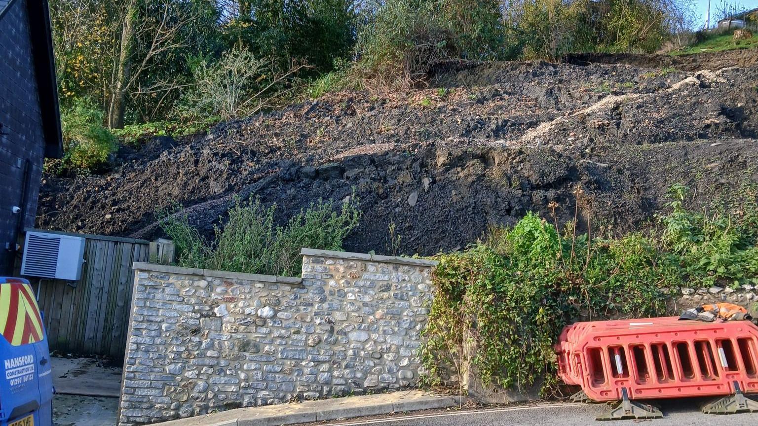 A collapsed bank next to a steep road. The collapse is behind a stone wall but the bare earth rises up behind it and appears to be about three times as high at the wall.