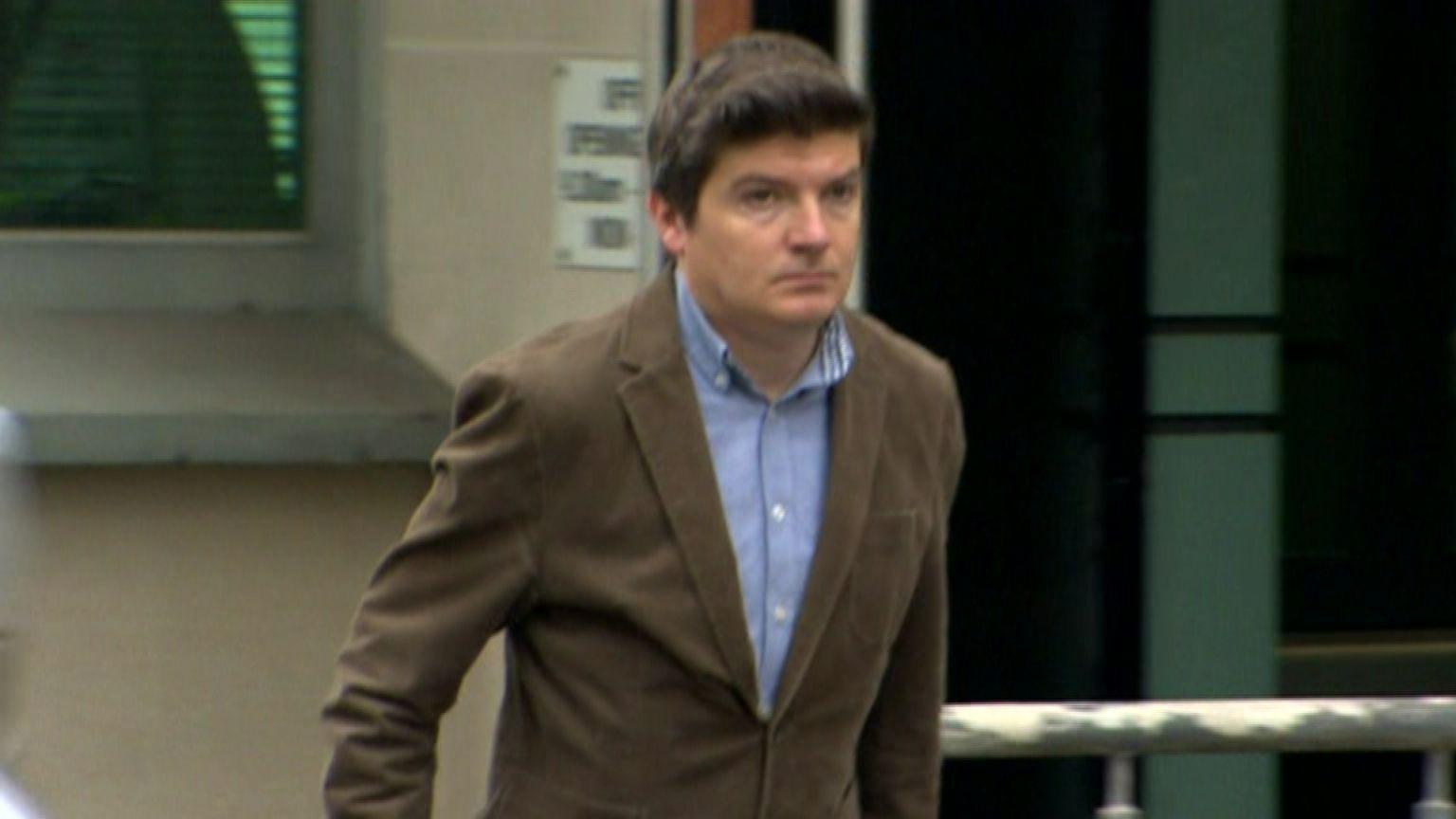 Michael McMonagle - a short dark-haired man wearing a brown, corduroy jacket and blue button-up collared shirt is walking. In the background is a white-bricked wall with a window, as well as a door.