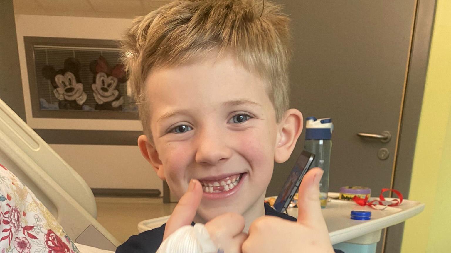 Lewis Danton sitting in a hospital bed in his room. He is smiling at the camera and is holding his thumbs up. 