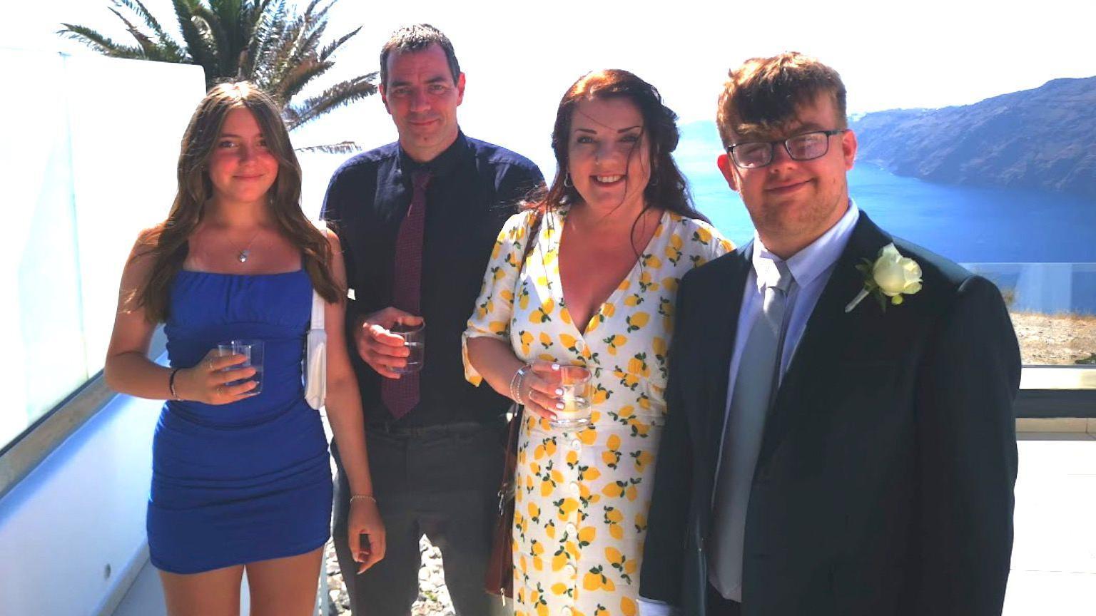 Chloe Longster stands on the far left smiling with a drink in her hand on holiday. She is smiling and wearing a blue bodycon dress.  Next to her, is her father wearing a black shirt and red tie. Her mother is in the middle, wearing a summer dress with lemons on it, she has the sides of her hair tied back. Chloe's brother, who is 19, is on the far right of the picture and has a white flower pinned to his grey suit. He is wearing glasses and has a light grey tie on