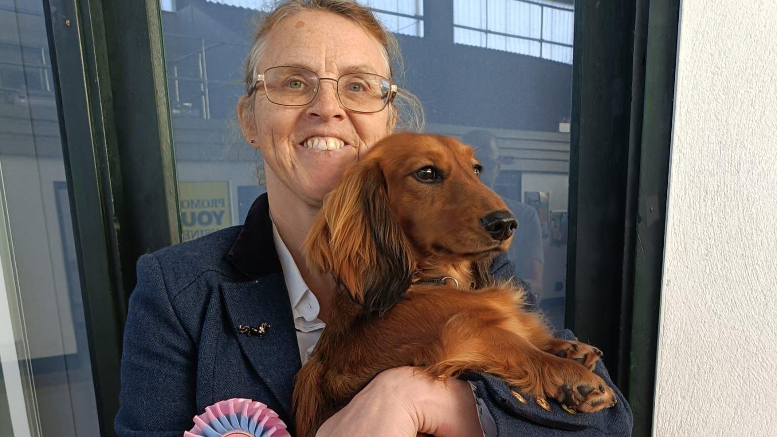 Susie Curteis-Lateo is holding her brown dachshund in her arms. Susie is wearing a blazer and glasses. She is smiling at the camera. Her dog has fluffy ears and is looking away from the camera.