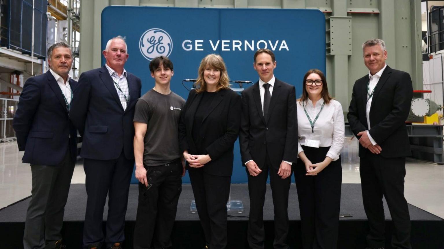 Seven people stand in a large factory, in front of a stage covered in a black material, which is in front of a blue sign which says "GE Vernova".