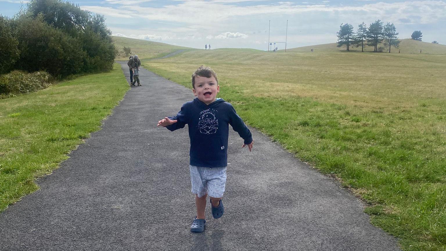 A young boy is smiling while running along a path in the countryside. He is wearing a blue hooded top and blue and white striped shorts and blue croc shoes. There are shrubs and trees in the distance. 