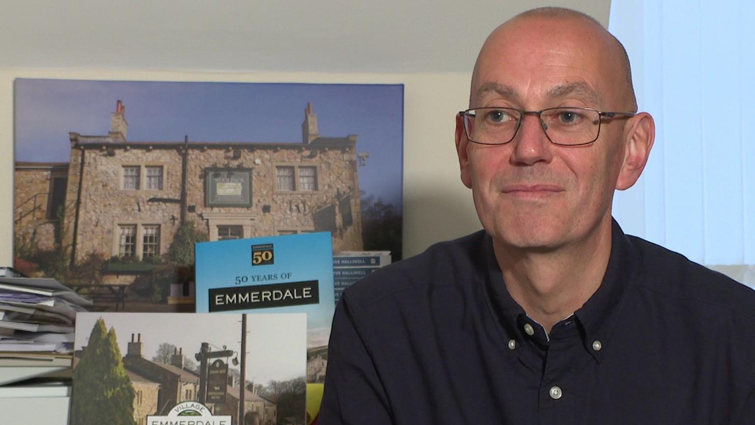 Steve Marshall has a bald head and is wearing a long-sleeve blue shirt. He has glasses on and behind him on a table is a picture of the Woolpack pub and a book celebrating 50 years of Emmerdale