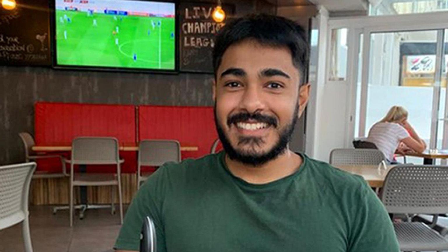 Anugrah Abraham, a man with black hair and facial hair and a dark green T-shirt, smiles directly into the camera. He is sat in a pub, with a TV displaying a football match behind his right shoulder.